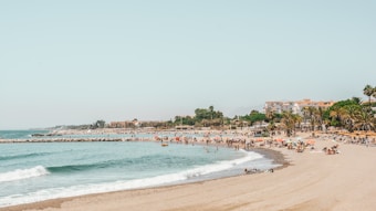 A sandy beach with numerous people enjoying the sun and sea. The coastline curves gently with waves lapping at the shore. Palm trees and buildings are visible in the background, suggesting a coastal resort area. Bright umbrellas and sun loungers dot the beach, creating a lively and bustling atmosphere.
