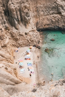 A small, secluded beach is nestled between high rocky cliffs with several colorful umbrellas and towels scattered on the sand. People are relaxing by the shore and enjoying the clear turquoise water. The atmosphere appears peaceful and leisurely, offering a sense of escape and tranquility.
