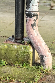 A weathered metal pipe with peeling paint stands next to a vertical black pipe. The pipes are mounted on a concrete block that is partially covered with moss and small patches of grass growing around it, set against a background of paving stones.
