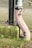 A weathered metal pipe with peeling paint stands next to a vertical black pipe. The pipes are mounted on a concrete block that is partially covered with moss and small patches of grass growing around it, set against a background of paving stones.