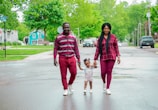 A family of three is walking hand in hand along a wet street. The parents are wearing coordinated maroon outfits and appear joyful as they assist their young child in walking. The background features a residential area with green trees and grass, creating a serene and pleasant atmosphere.