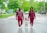 A family of three is walking hand in hand along a wet street. The parents are wearing coordinated maroon outfits and appear joyful as they assist their young child in walking. The background features a residential area with green trees and grass, creating a serene and pleasant atmosphere.