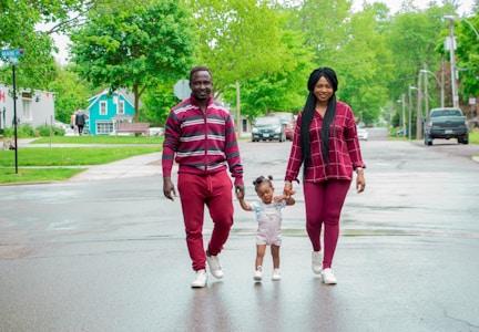 A family of three is walking hand in hand along a wet street. The parents are wearing coordinated maroon outfits and appear joyful as they assist their young child in walking. The background features a residential area with green trees and grass, creating a serene and pleasant atmosphere.