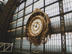 An ornate, antique clock with Roman numerals is prominently displayed in a grand hall. The clock features intricate gold detailing and is set against a backdrop of large, industrial-style windows with geometric patterns. The architecture suggests an elegant, historical setting.