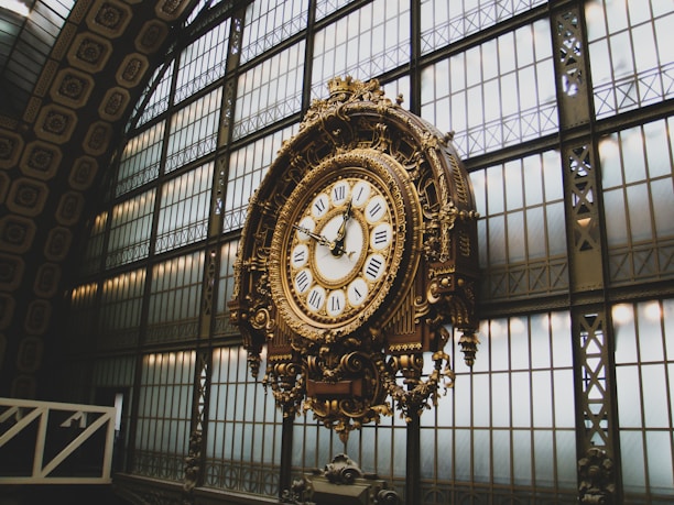 An ornate, antique clock with Roman numerals is prominently displayed in a grand hall. The clock features intricate gold detailing and is set against a backdrop of large, industrial-style windows with geometric patterns. The architecture suggests an elegant, historical setting.
