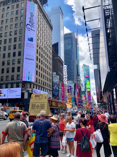 A bustling urban street scene with a diverse crowd of people walking along a busy avenue filled with tall buildings. Brightly lit advertisements and billboards cover the facades, and a big tour bus is seen in the foreground with its passengers. The sky is partly cloudy, adding a mix of shadows and light to the scene.