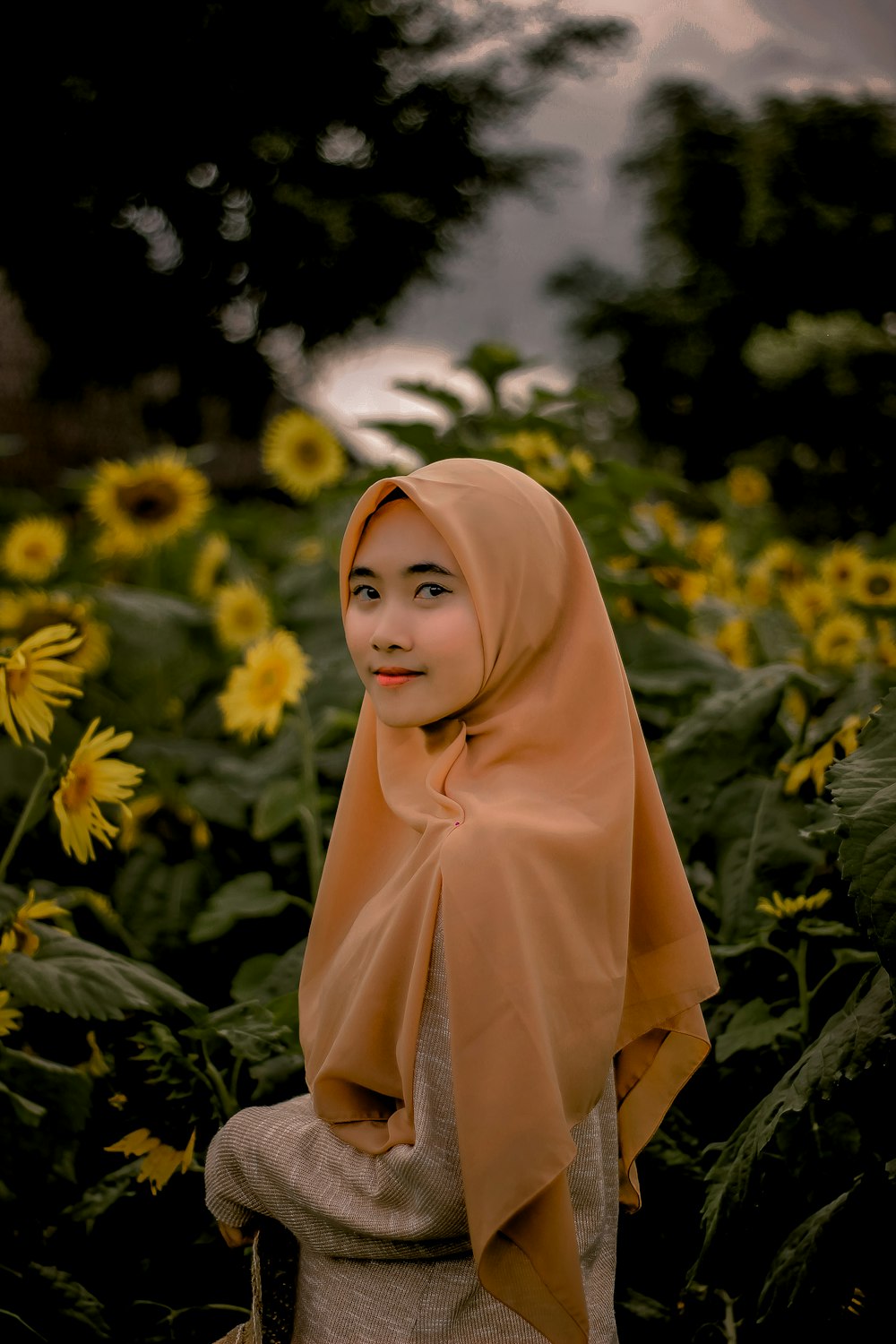 woman in brown abaya dress and orange hijab standing in blooming sunflower garden