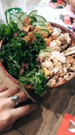 A wooden bowl filled with a variety of fresh ingredients including kale, sliced cucumbers, seaweed, and a creamy mixture likely containing tofu or a similar protein. A person with a decorative ring on their finger holds the edge of the bowl on a wooden table. A Starbucks cup is partially visible in the background along with a menu featuring food images.