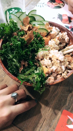 A wooden bowl filled with a variety of fresh ingredients including kale, sliced cucumbers, seaweed, and a creamy mixture likely containing tofu or a similar protein. A person with a decorative ring on their finger holds the edge of the bowl on a wooden table. A Starbucks cup is partially visible in the background along with a menu featuring food images.