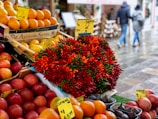An assorted display of various fruits and vegetables at a market. Boxes of oranges, nectarines, and grapefruits are arranged neatly. A large bouquet of colorful red and green chili peppers is prominently placed in the center. Handwritten price tags are visible, suggesting a lively market atmosphere. In the blurred background, people are walking along a wet pavement, indicating a recent rainfall.