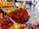 An assorted display of various fruits and vegetables at a market. Boxes of oranges, nectarines, and grapefruits are arranged neatly. A large bouquet of colorful red and green chili peppers is prominently placed in the center. Handwritten price tags are visible, suggesting a lively market atmosphere. In the blurred background, people are walking along a wet pavement, indicating a recent rainfall.