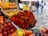 An assorted display of various fruits and vegetables at a market. Boxes of oranges, nectarines, and grapefruits are arranged neatly. A large bouquet of colorful red and green chili peppers is prominently placed in the center. Handwritten price tags are visible, suggesting a lively market atmosphere. In the blurred background, people are walking along a wet pavement, indicating a recent rainfall.