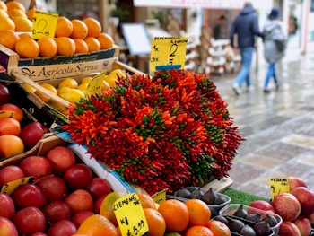 An assorted display of various fruits and vegetables at a market. Boxes of oranges, nectarines, and grapefruits are arranged neatly. A large bouquet of colorful red and green chili peppers is prominently placed in the center. Handwritten price tags are visible, suggesting a lively market atmosphere. In the blurred background, people are walking along a wet pavement, indicating a recent rainfall.