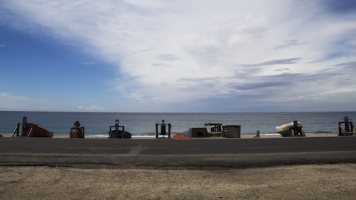 A coastal scene with a calm ocean in the background and a partially cloudy sky. Along the shoreline are several pieces of equipment or structures aligned in a row. The foreground features an asphalt road and sandy ground.