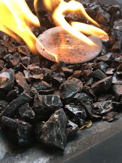 A close-up view of a fire pit with a circular metal surface surrounded by dark, jagged stones. Bright, orange flames are rising above the stones, casting a warm light and slight reflections on the surrounding materials.
