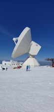 A large satellite dish is positioned on a snow-covered terrain under a clear blue sky. Several people are walking nearby, and some buildings and structures are visible in the background.
