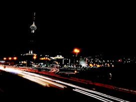 A cityscape captured at night with a skyline featuring a tall tower illuminated by lights. The foreground displays streaks of light from vehicles on a highway, suggesting long exposure photography.