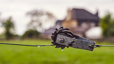 A close-up view of a metal ratchet tensioner attached to a wire fence. The blurred background reveals a green field, some trees, and an out-of-focus building. The scene appears to be outdoors, possibly in a rural setting, after a recent rainfall as indicated by a drop of water hanging from the wire.