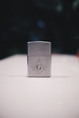 A metallic rectangular lighter sits on a white surface. The lighter is adorned with a geometric emblem, featuring a capital 'G' centered between a compass and square, symbols often associated with Freemasonry. The background is dark and blurred, drawing focus to the foreground object.