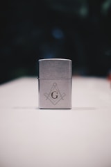 A metallic rectangular lighter sits on a white surface. The lighter is adorned with a geometric emblem, featuring a capital 'G' centered between a compass and square, symbols often associated with Freemasonry. The background is dark and blurred, drawing focus to the foreground object.