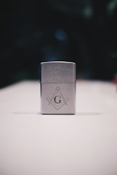 A metallic rectangular lighter sits on a white surface. The lighter is adorned with a geometric emblem, featuring a capital 'G' centered between a compass and square, symbols often associated with Freemasonry. The background is dark and blurred, drawing focus to the foreground object.