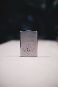A metallic rectangular lighter sits on a white surface. The lighter is adorned with a geometric emblem, featuring a capital 'G' centered between a compass and square, symbols often associated with Freemasonry. The background is dark and blurred, drawing focus to the foreground object.