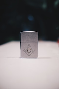 A metallic rectangular lighter sits on a white surface. The lighter is adorned with a geometric emblem, featuring a capital 'G' centered between a compass and square, symbols often associated with Freemasonry. The background is dark and blurred, drawing focus to the foreground object.