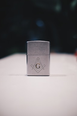 A metallic rectangular lighter sits on a white surface. The lighter is adorned with a geometric emblem, featuring a capital 'G' centered between a compass and square, symbols often associated with Freemasonry. The background is dark and blurred, drawing focus to the foreground object.