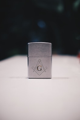 A metallic rectangular lighter sits on a white surface. The lighter is adorned with a geometric emblem, featuring a capital 'G' centered between a compass and square, symbols often associated with Freemasonry. The background is dark and blurred, drawing focus to the foreground object.