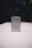 A metallic rectangular lighter sits on a white surface. The lighter is adorned with a geometric emblem, featuring a capital 'G' centered between a compass and square, symbols often associated with Freemasonry. The background is dark and blurred, drawing focus to the foreground object.