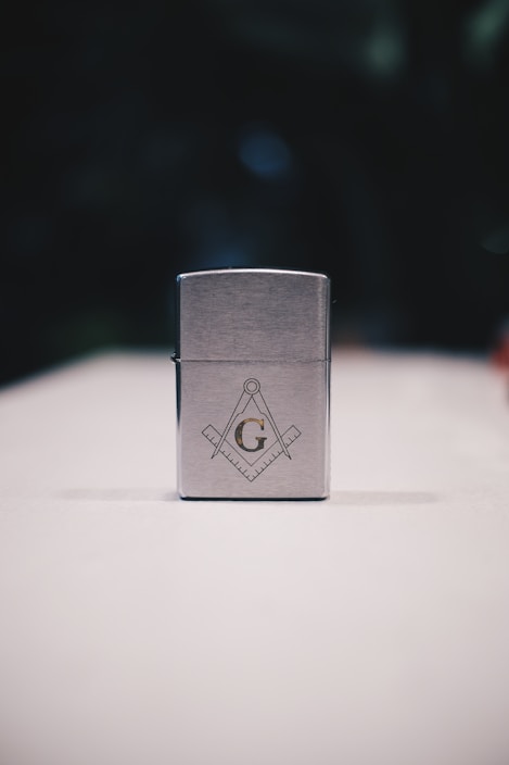 A metallic rectangular lighter sits on a white surface. The lighter is adorned with a geometric emblem, featuring a capital 'G' centered between a compass and square, symbols often associated with Freemasonry. The background is dark and blurred, drawing focus to the foreground object.