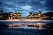 A beachside evening event is set up with tables and chairs under strings of warm lights. The gathering is well attended, with guests mingling and dining. Palm trees and buildings form the backdrop under a cloudy blue sky, with the scene beautifully reflected in a nearby water pool.