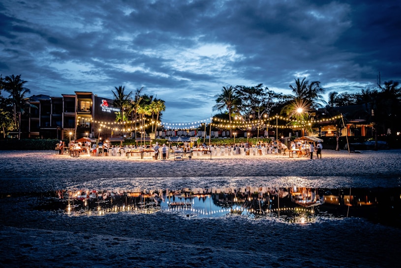 A beachside evening event is set up with tables and chairs under strings of warm lights. The gathering is well attended, with guests mingling and dining. Palm trees and buildings form the backdrop under a cloudy blue sky, with the scene beautifully reflected in a nearby water pool.
