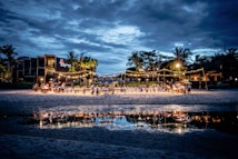 A beachside evening event is set up with tables and chairs under strings of warm lights. The gathering is well attended, with guests mingling and dining. Palm trees and buildings form the backdrop under a cloudy blue sky, with the scene beautifully reflected in a nearby water pool.