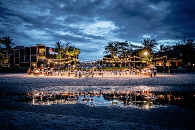 A beachside evening event is set up with tables and chairs under strings of warm lights. The gathering is well attended, with guests mingling and dining. Palm trees and buildings form the backdrop under a cloudy blue sky, with the scene beautifully reflected in a nearby water pool.