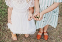 Two children dressed in vintage-style clothing stand together on grass. One child wears a white dress with lace detail, while the other wears a green and white striped dress with buttons. They hold small objects in their hands, one with a black object and the other with green leaves.