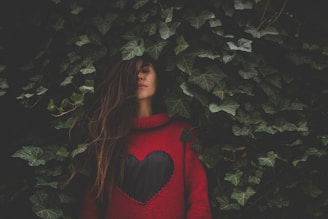 A young woman with long hair stands amidst dense green ivy leaves. She wears a red sweater with a large black heart design on the front. Her eyes are closed, and she appears contemplative, partially obscured by the foliage around her.