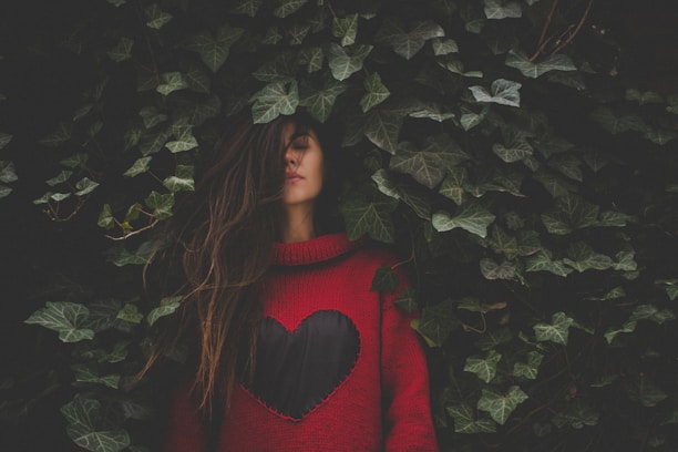 A young woman with long hair stands amidst dense green ivy leaves. She wears a red sweater with a large black heart design on the front. Her eyes are closed, and she appears contemplative, partially obscured by the foliage around her.
