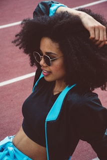 A stylish woman with voluminous curly hair wearing sunglasses and a sporty outfit that includes a black crop top and a jacket with blue accents is seen relaxing on a track field.
