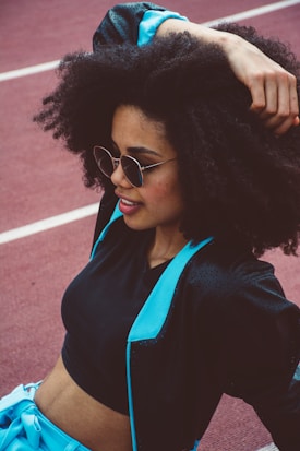 A stylish woman with voluminous curly hair wearing sunglasses and a sporty outfit that includes a black crop top and a jacket with blue accents is seen relaxing on a track field.