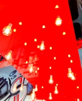 A modern lighting installation features numerous exposed light bulbs hanging from a ceiling with a vibrant, glossy red background. The bulbs emit a warm yellow glow. Below, a partial view of a wall with abstract black and white designs is visible.