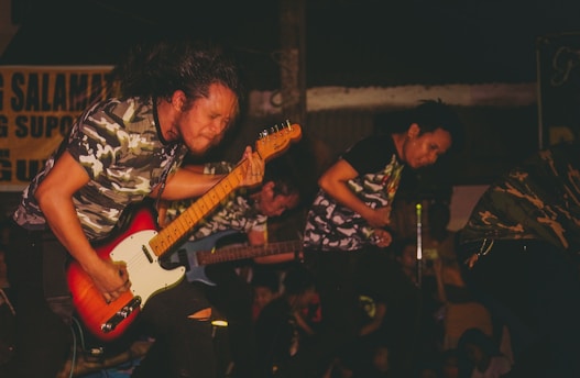 A group of musicians passionately performing on stage. The focus is on a guitarist with long hair, wearing a camo t-shirt, energetically playing an electric guitar. Other band members are partially visible, wearing similar camouflage attire.