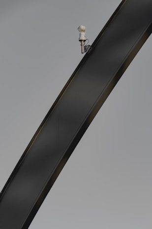 A sleek, curved metal structure with a mounted security camera positioned near the top against a clear background. The image highlights the curvature and texture of the metal beam, and the camera is small yet distinct in its placement.