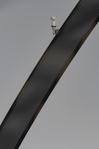 A sleek, curved metal structure with a mounted security camera positioned near the top against a clear background. The image highlights the curvature and texture of the metal beam, and the camera is small yet distinct in its placement.