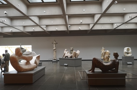 A modern art gallery with several abstract sculptures displayed on pedestals. The sculptures vary in shape and material, occupying a spacious, well-lit room with a geometric ceiling. Visitors are seen observing the artworks.