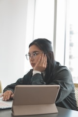 A person sitting at a table looking at a laptop or tablet with a thoughtful expression. Natural light filters in through large windows, creating a bright and calm environment. The person wears glasses and a dark top, supporting their chin with a hand while using the other hand on the laptop.