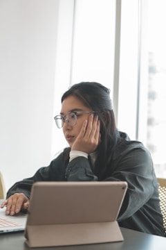 A person sitting at a table looking at a laptop or tablet with a thoughtful expression. Natural light filters in through large windows, creating a bright and calm environment. The person wears glasses and a dark top, supporting their chin with a hand while using the other hand on the laptop.