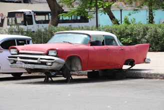 An old, red vintage car is parked on the street without wheels, supported by blocks. The car has a white roof and is in a state of disrepair, missing headlights and showing signs of rust. In the background, there is greenery and another parked vehicle partially visible. A white bus can be seen driving past on the road behind the cars.