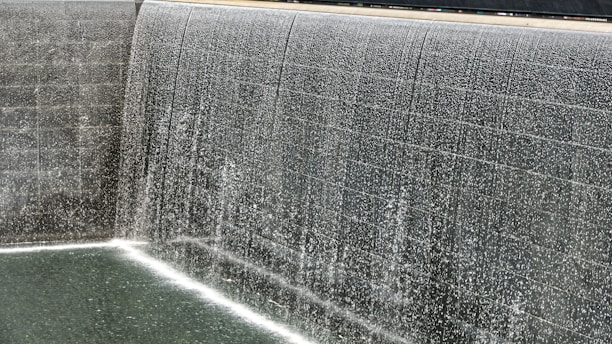A cascading waterfall flows over dark tiled walls into a calm pool below. The water creates a shimmering effect as it falls, with droplets and splashes captured mid-motion. The texture of the tile provides a contrasting backdrop to the fluid movement of the water.