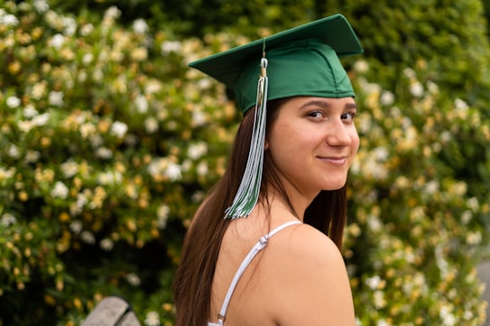 A young person wearing a green graduation cap with a tassel is looking over their shoulder, smiling slightly. They are standing in front of a lush, green bush with small white flowers. The person's long hair flows down their back and they are wearing a light-colored, sleeveless top.